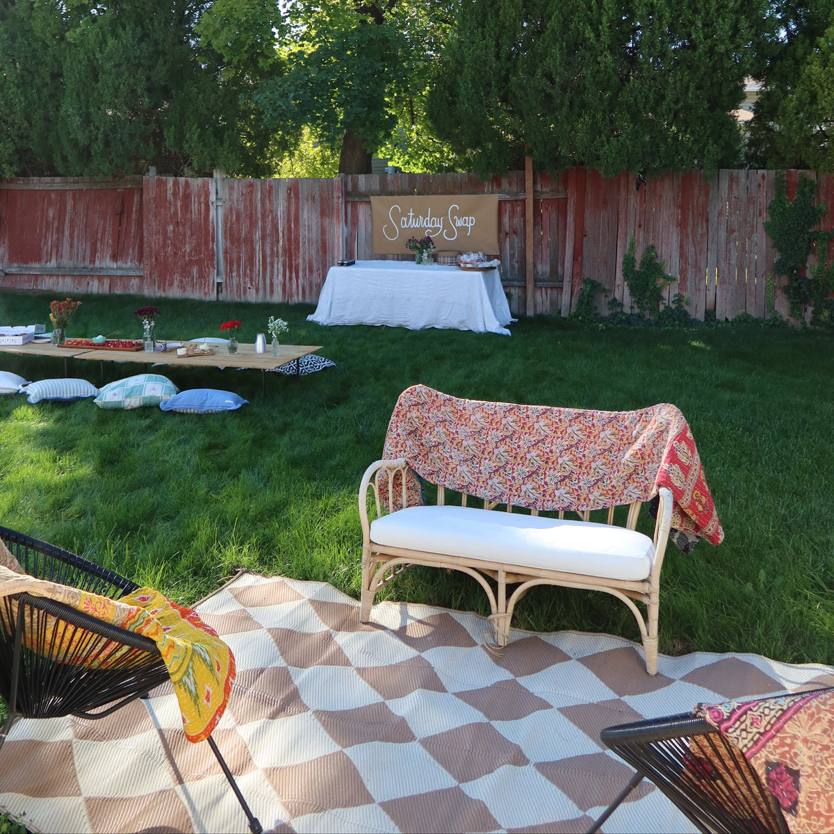 Outdoor backyard event setup with a white lounge chair draped in a floral throw blanket kantha quilt, a checkered rug, a wooden fence, and a decorated table in the background.